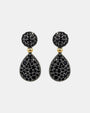 Pareja de pendientes articulados Bohemme con botón y lágrima en pavé de espinelas negras sobre fondo gris claro.