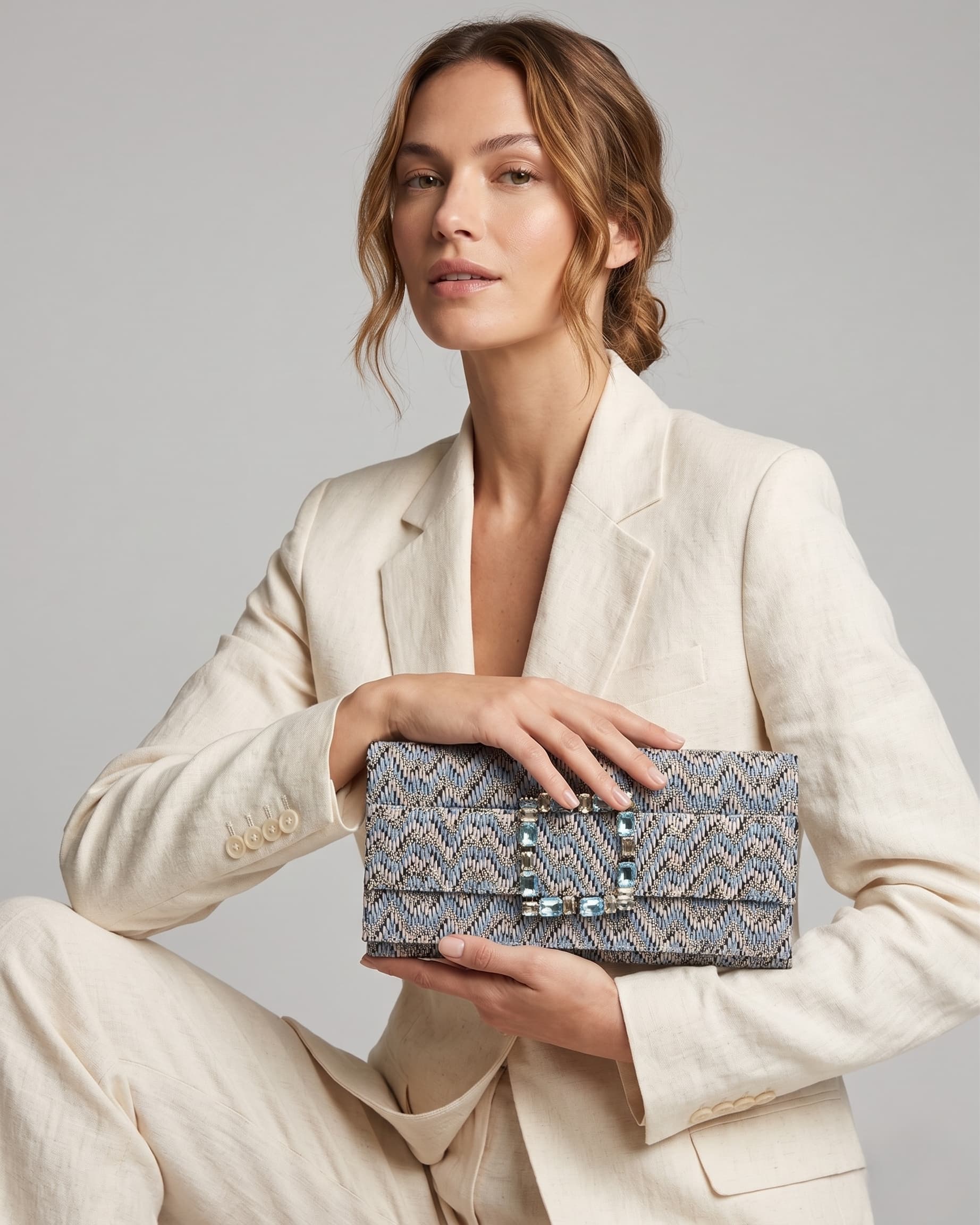 modelo sentada posando con un traje de chaqueta blanco de lino mientras sujeta la cartera de Emanuela Caruso elegantemente con ambas manos sobre sus piernas.