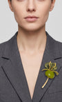 Vista frontal del maxibroche floral verde de Ciléa Paris prendido en la solapa de una chaqueta de traje gris oscuro, detallando su escala y volumen tridimensional.