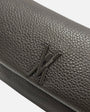 primer plano macro del Bolso Bandolera Cacao de Plinio Visonà, permitiendo apreciar la textura de la piel de grano y el monograma metálico.
