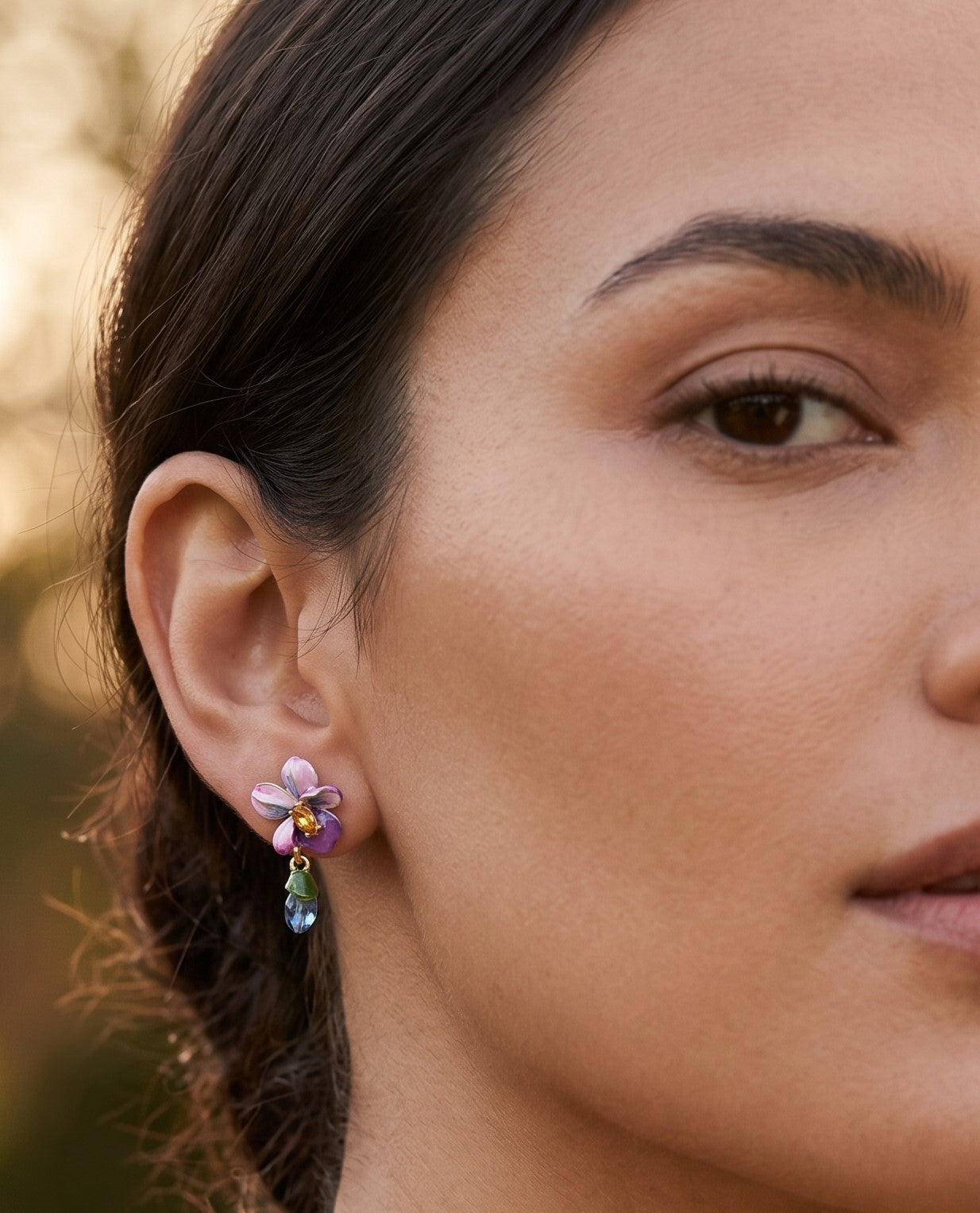 Modelo luciendo el pendiente asimétrico Les Néréides Violette Sauvage con flor de orquídea esmaltada y gota azul colgando sobre el lóbulo.