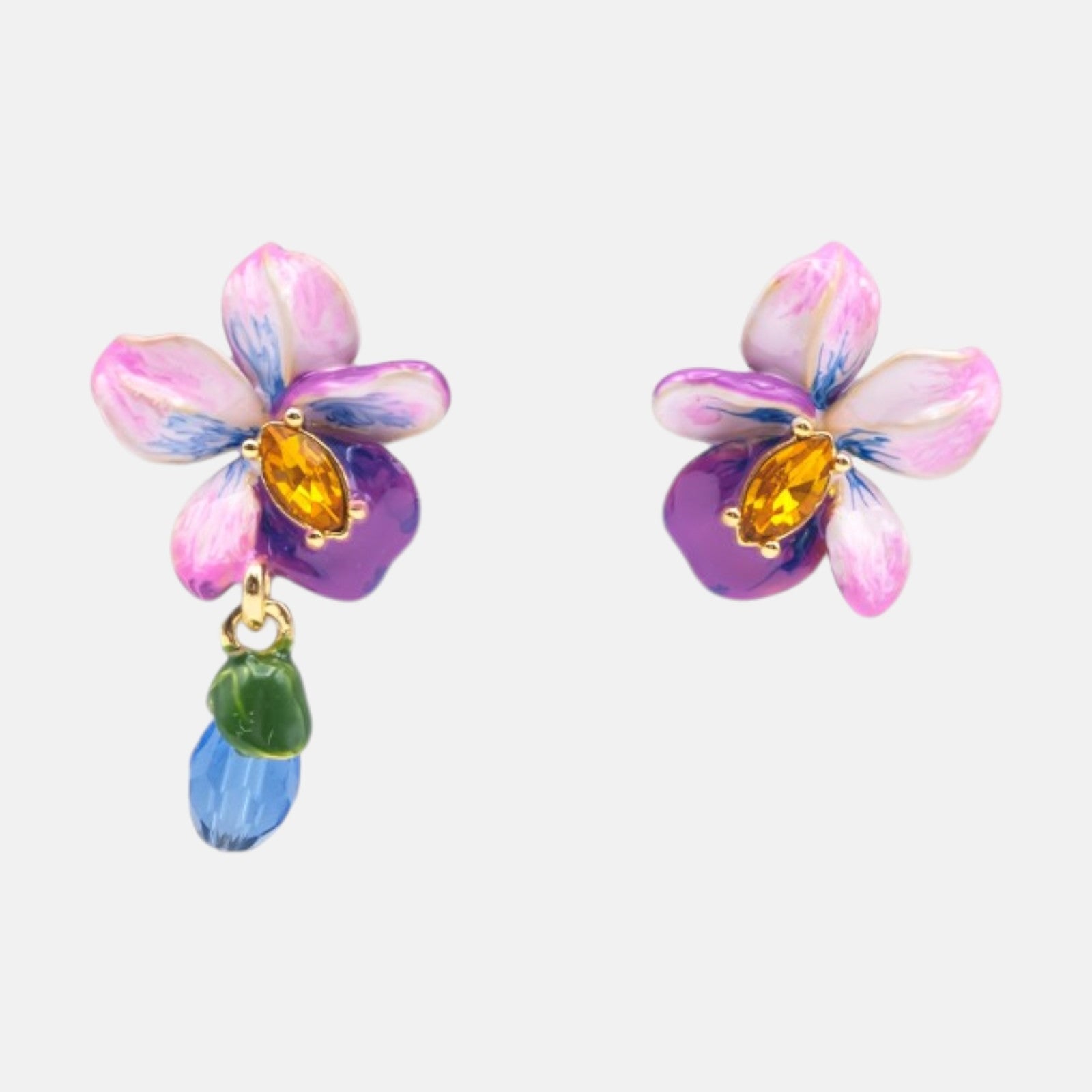 Par de pendientes asimétricos Les Néréides Violette Sauvage con orquídeas esmaltadas, cristales ámbar y gota de cristal azul sobre fondo neutro.