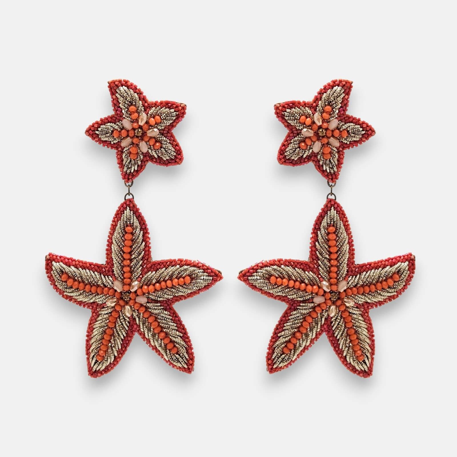 Par de pendientes maxi Océano Vivo con diseño de estrella de mar bordados en pedrería coral y dorado sobre fondo neutro.