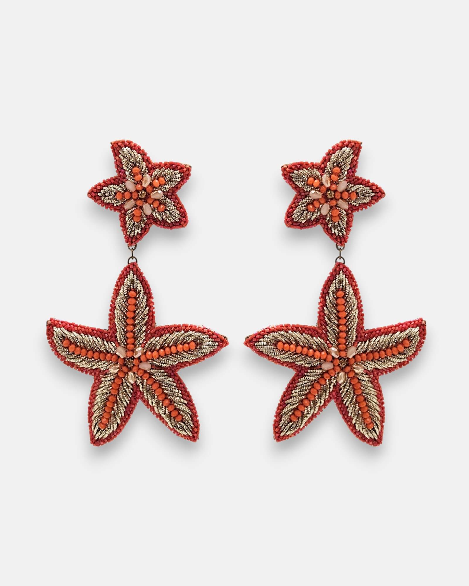 Par de pendientes maxi Océano Vivo con diseño de estrella de mar bordados en pedrería coral y dorado sobre fondo neutro.