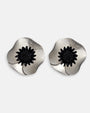par de pendientes de clip con forma de flor de amapola de Ciléa Paris, detallando sus pétalos de resina satinada en tono plata metalizada y su centro negro estrellado.