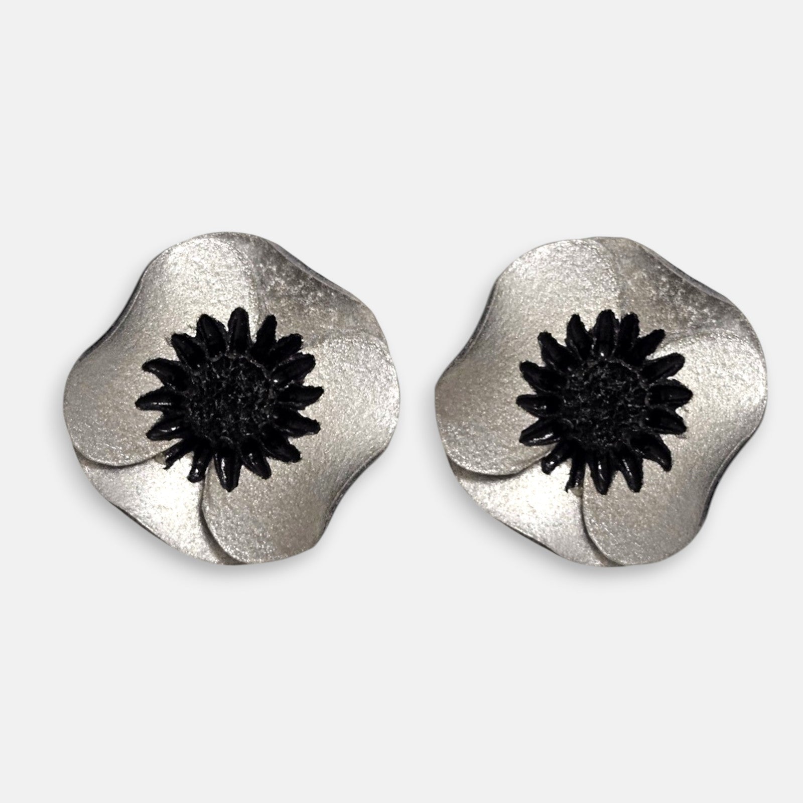 par de pendientes de clip con forma de flor de amapola de Ciléa Paris, detallando sus pétalos de resina satinada en tono plata metalizada y su centro negro estrellado.
