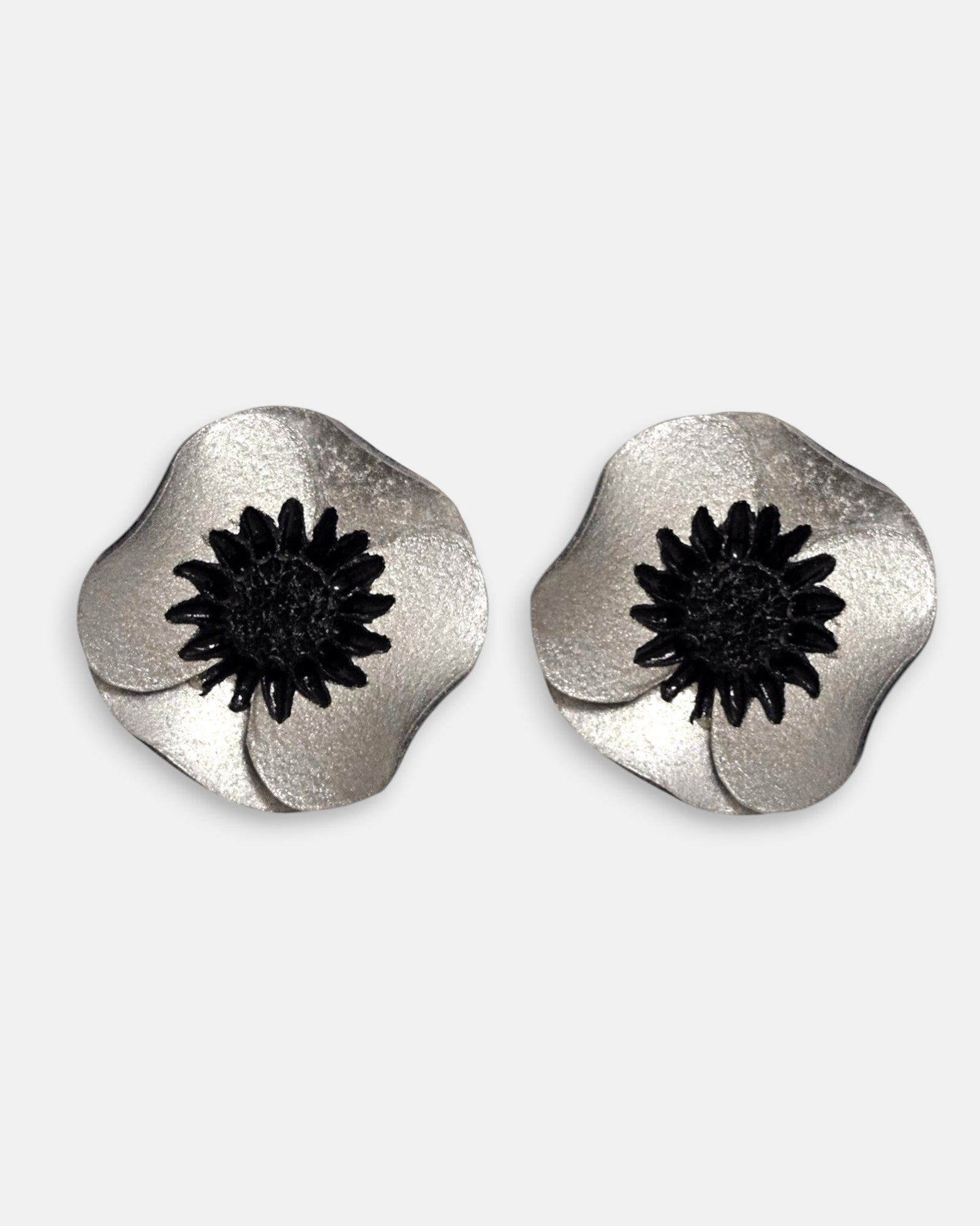 par de pendientes de clip con forma de flor de amapola de Ciléa Paris, detallando sus pétalos de resina satinada en tono plata metalizada y su centro negro estrellado.