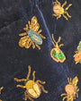 primer plano macro del tejido de terciopelo azul noche, permitiendo apreciar la precisión del bordado a mano de los escarabajos y abejas en cuentas doradas y verdes.