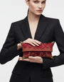 Bolso clutch de mano tipo sobre en tejido jacquard granate con bordado de rayas ocres y esquineras metálicas doradas, lucido con americana negra.
