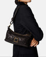 modelo luciendo el bolso Camilla colgado al hombro sobre un traje oscuro, apreciando su tamaño compacto y su estilo sobrio.