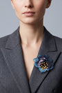 modelo de frente luciendo el broche maxi de loto azul prendido en la solapa de una americana de sastre gris oscuro, mostrando su escala sofisticada y elegante.