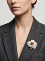 modelo luciendo el broche maxi de loto blanco prendido en la solapa de una chaqueta de sastre gris oscuro, apreciando su escala y su sofisticado diseño.