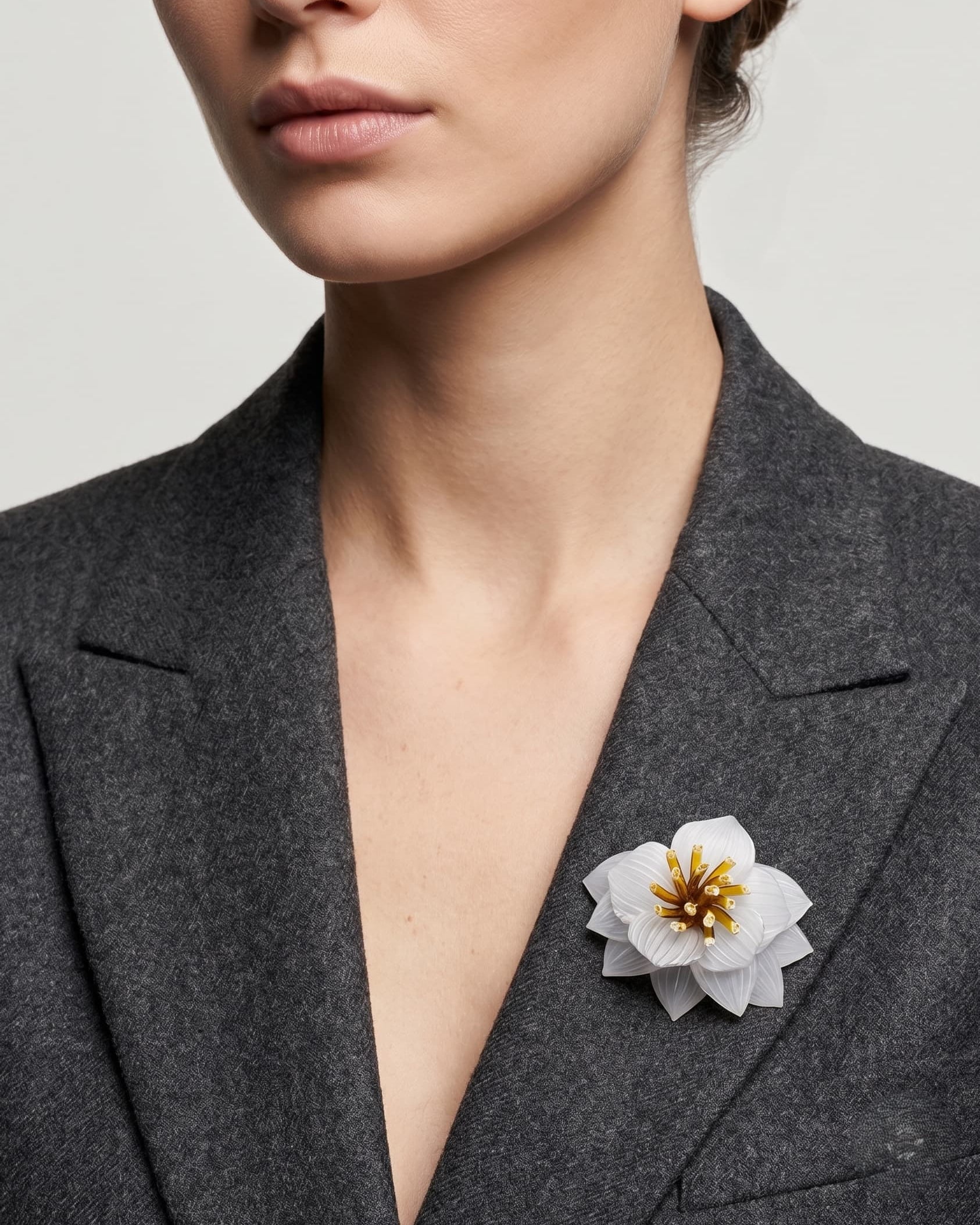 modelo luciendo el broche maxi de loto blanco prendido en la solapa de una chaqueta de sastre gris oscuro, apreciando su escala y su sofisticado diseño.