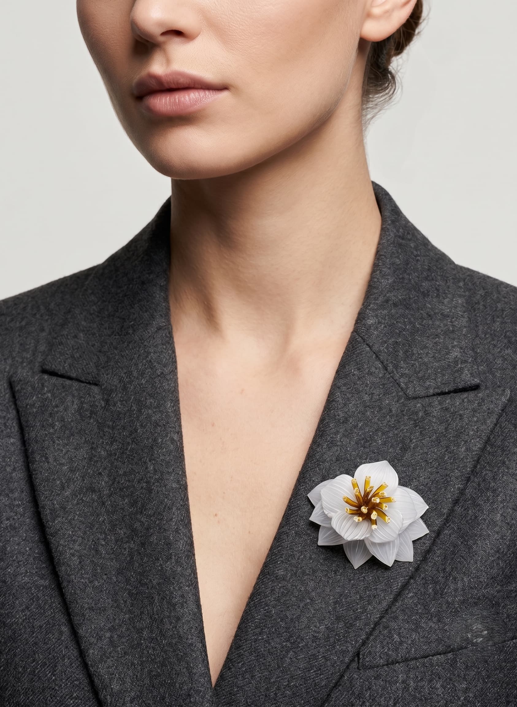 modelo luciendo el broche maxi de loto blanco prendido en la solapa de una chaqueta de sastre gris oscuro, apreciando su escala y su sofisticado diseño.