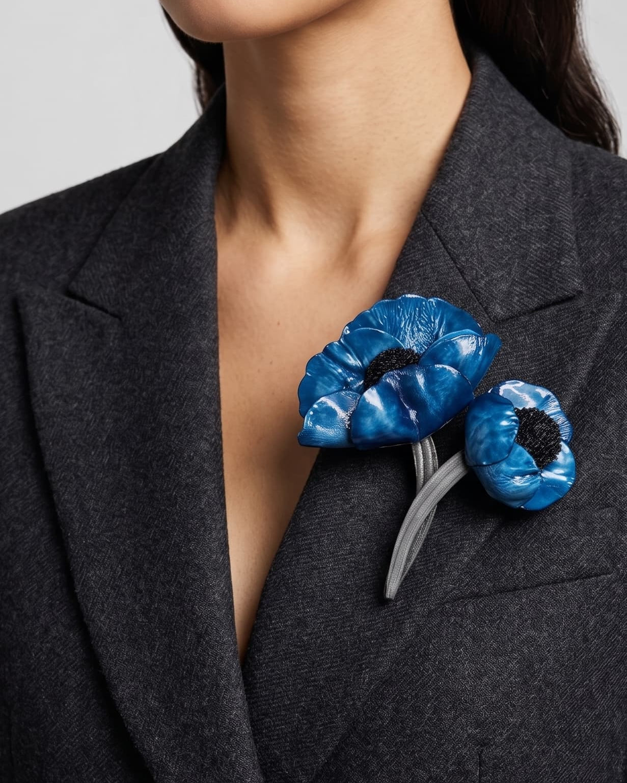 modelo luciendo el broche maxi de doble amapola azul prendido en la solapa de una chaqueta de sastre gris oscuro, mostrando su escala maximalista y elegante.