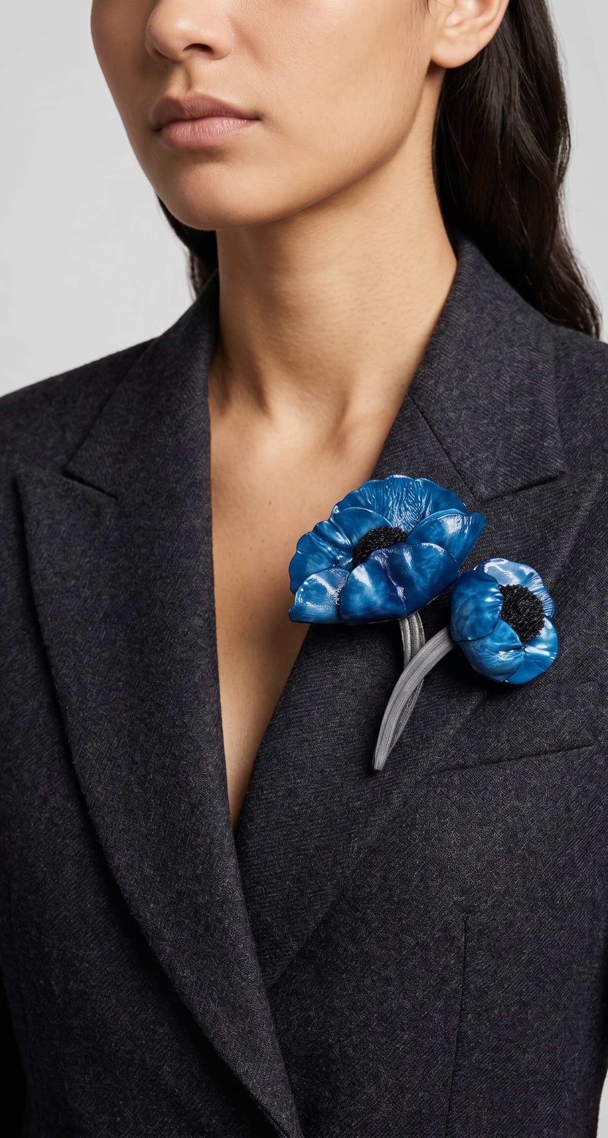 modelo luciendo el broche maxi de doble amapola azul prendido en la solapa de una chaqueta de sastre gris oscuro, mostrando su escala maximalista y elegante.