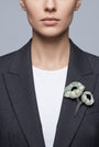 modelo de frente luciendo el broche maxi de doble amapola verde menta prendido en la solapa de una chaqueta de sastre gris oscuro, mostrando su escala maximalista y elegante.