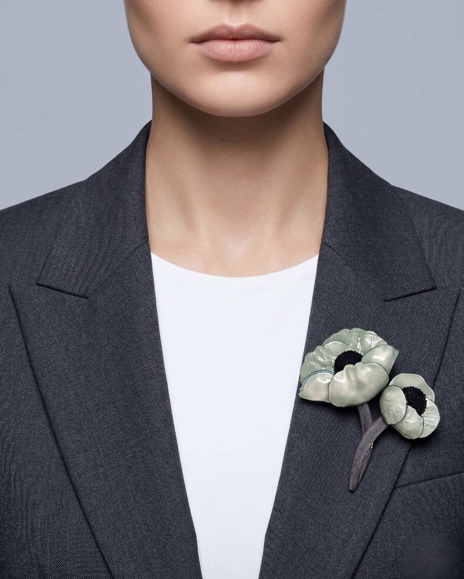 modelo de frente luciendo el broche maxi de doble amapola verde menta prendido en la solapa de una chaqueta de sastre gris oscuro, mostrando su escala maximalista y elegante.