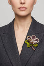 modelo luciendo el broche maxi de amapola violeta prendido en la solapa de una chaqueta de sastre gris oscuro, mostrando su escala elegante y volumetría.