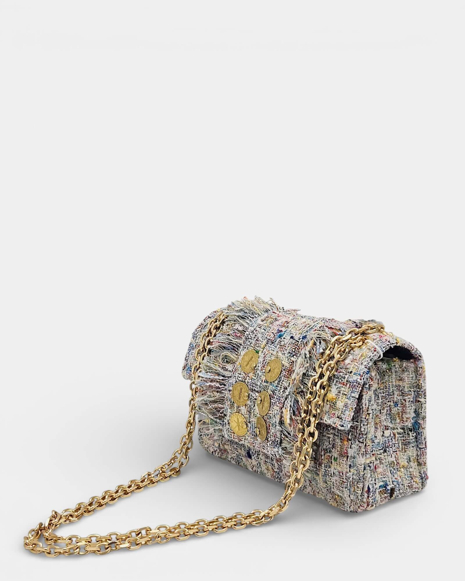 Vista en ángulo de bolso bandolera de tweed color crema mostrando la doble cadena metálica dorada y textura de flecos.