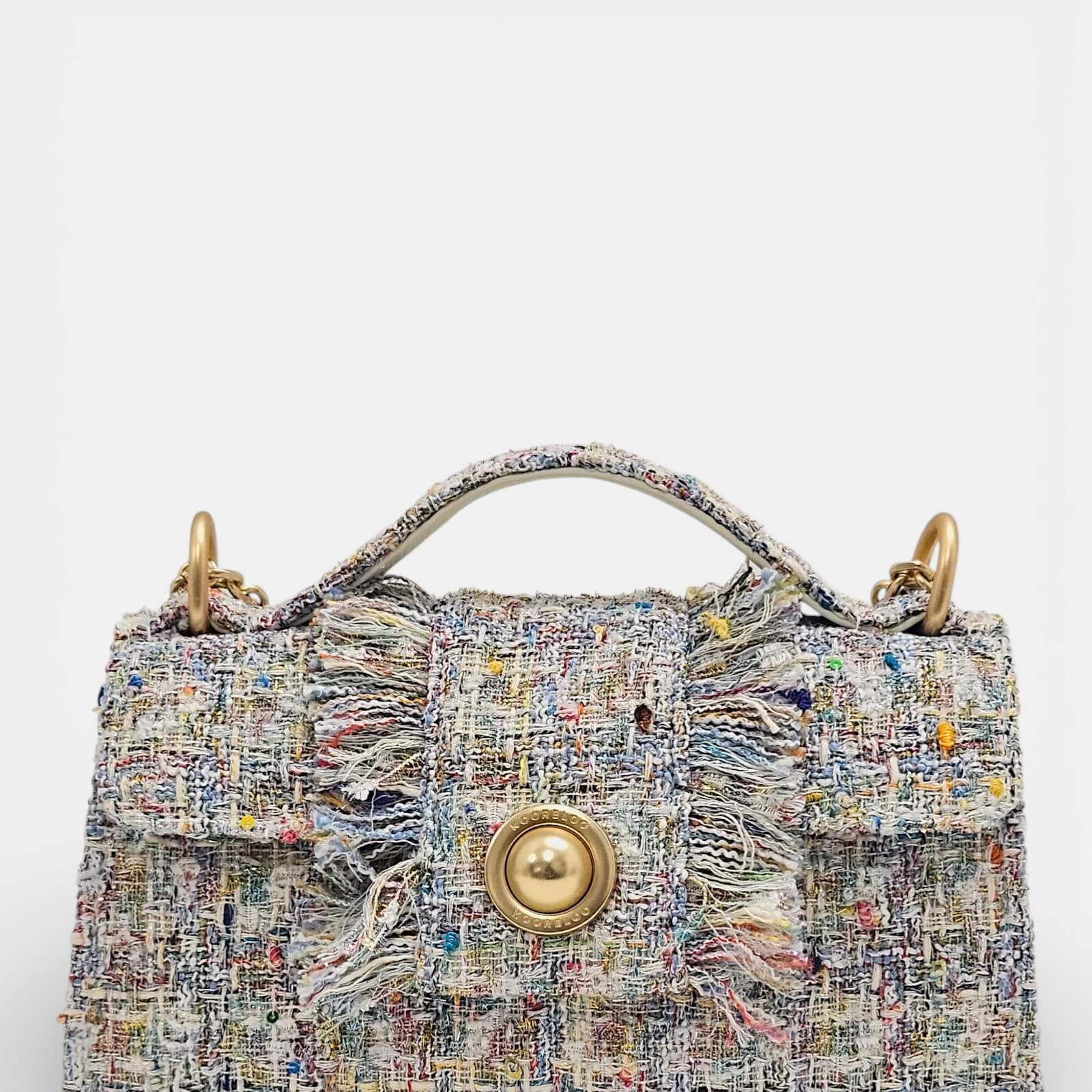 Bolso de mano en tweed color beige y pastel con una gran moneda dorada central, asa superior rígida y bordes con flecos.