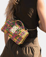 Mujer de espaldas luciendo un bolso bandolera de tweed en tonos rosa, verde y amarillo con cadena dorada y logo circular metálico.