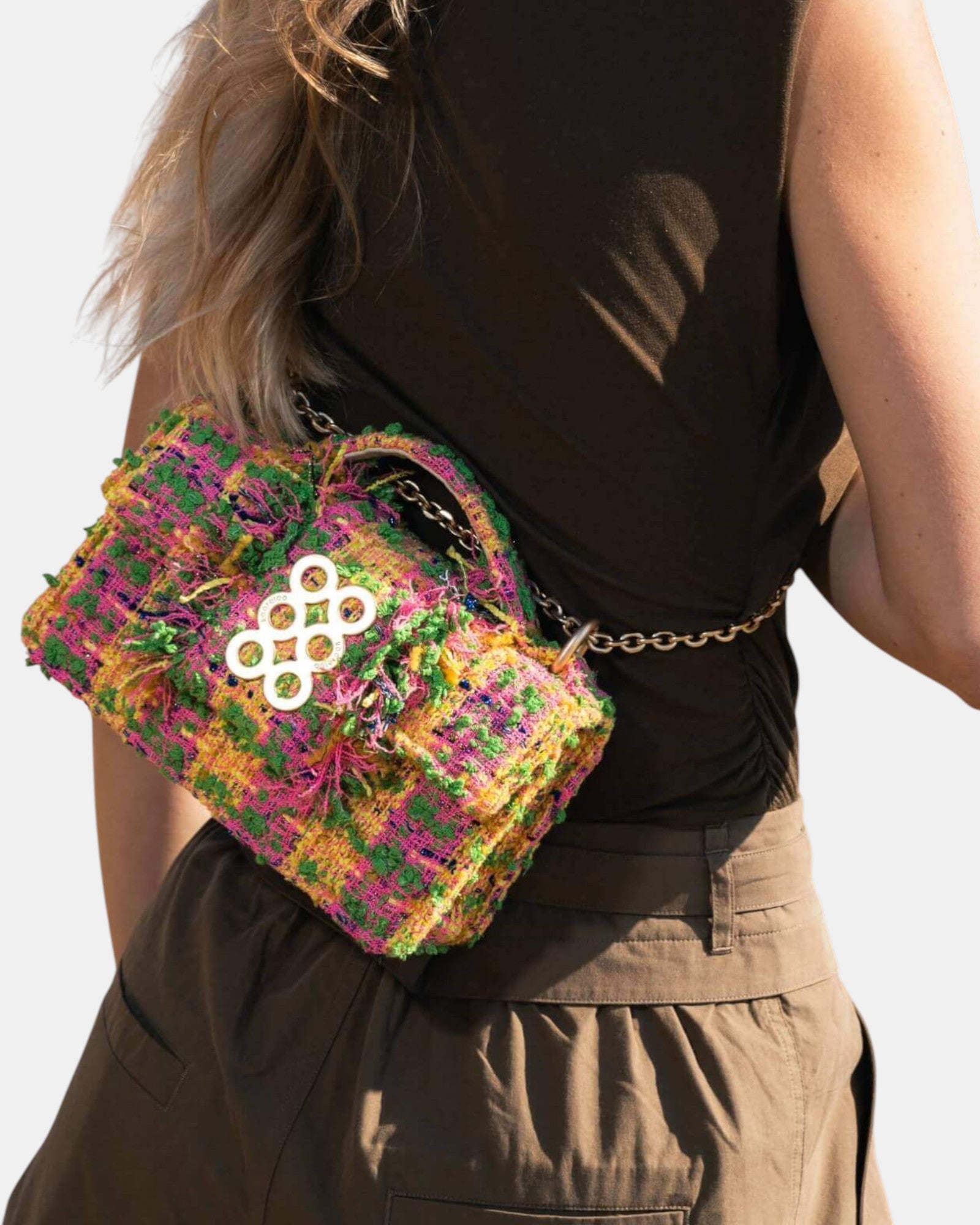 Mujer de espaldas luciendo un bolso bandolera de tweed en tonos rosa, verde y amarillo con cadena dorada y logo circular metálico.