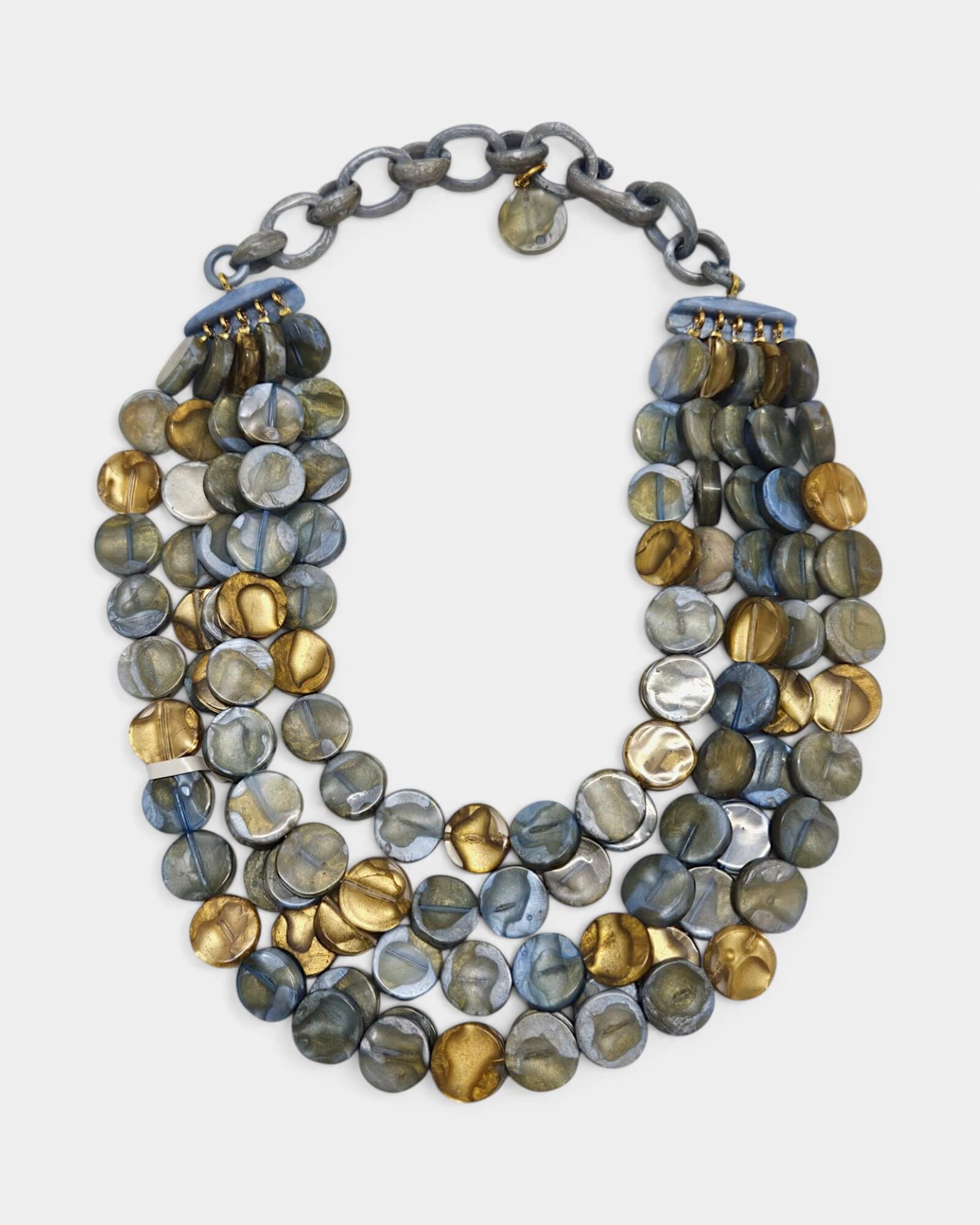 Collar de cuatro hileras Lisa de Dominique Denaive con cuentas circulares en azul, gris nacarado y oro sobre fondo blanco.