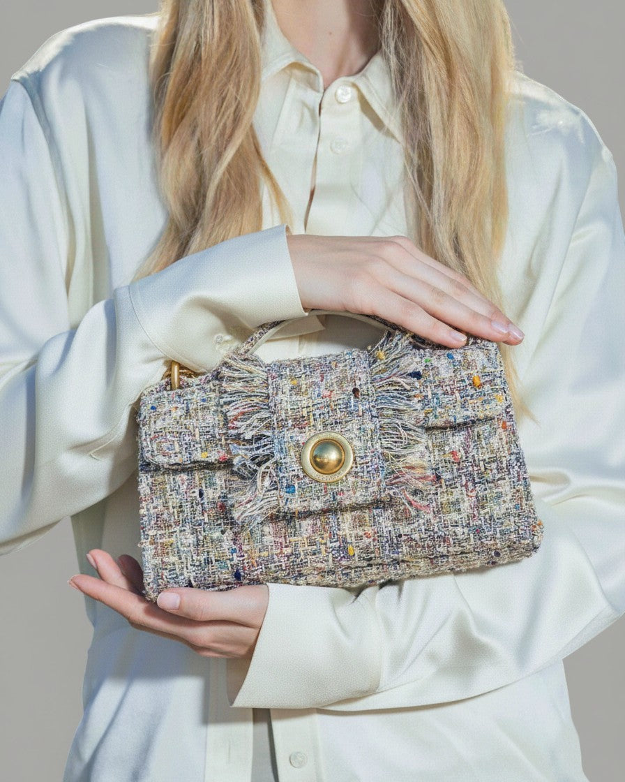 Modelo sosteniendo un bolso de mano de tweed con moneda dorada central y flecos, combinado con una camisa de seda color crema.