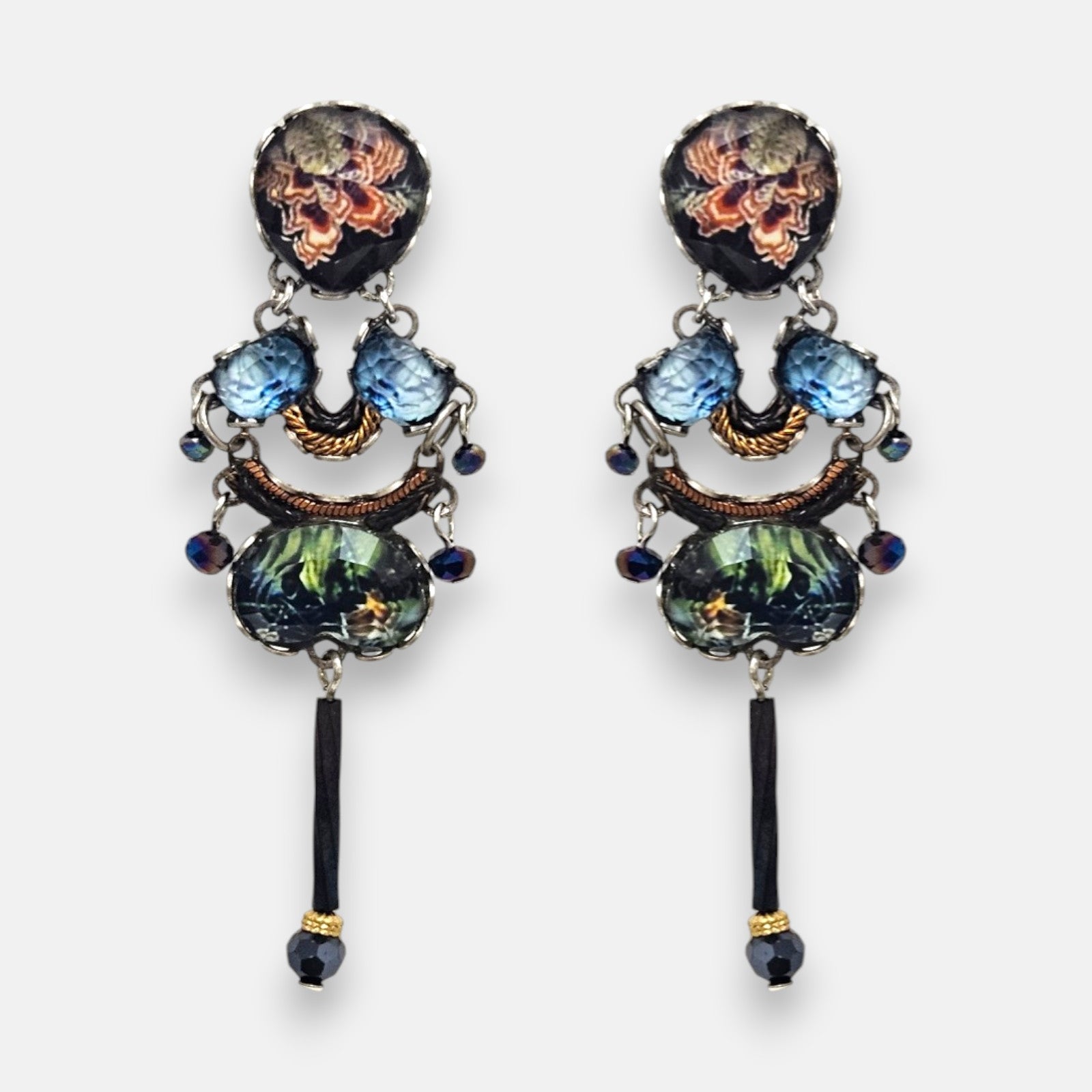 par de pendientes expuesto de frente sobre un fondo neutro, detallando su silueta simétrica con flores coral en el botón, cristales celestes y la aguja negra inferior.