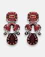 par de pendientes de frente sobre un fondo gris claro, destacando su silueta simétrica de estilo barroco con cristales rosas, detalles textiles arcilla y la lágrima central granate.