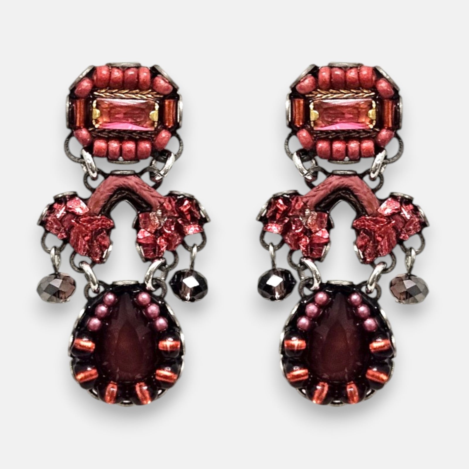 par de pendientes de frente sobre un fondo gris claro, destacando su silueta simétrica de estilo barroco con cristales rosas, detalles textiles arcilla y la lágrima central granate.