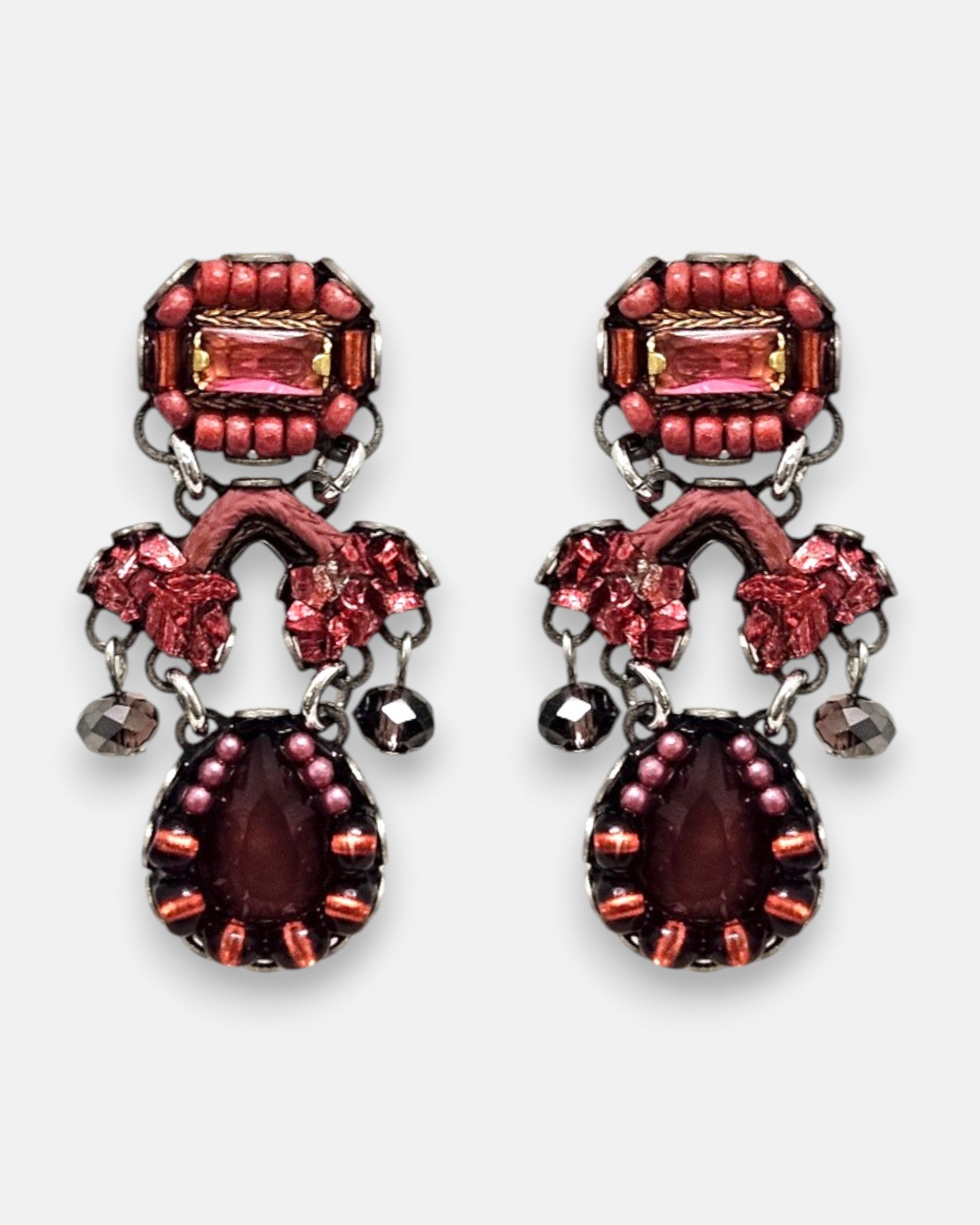 par de pendientes de frente sobre un fondo gris claro, destacando su silueta simétrica de estilo barroco con cristales rosas, detalles textiles arcilla y la lágrima central granate.
