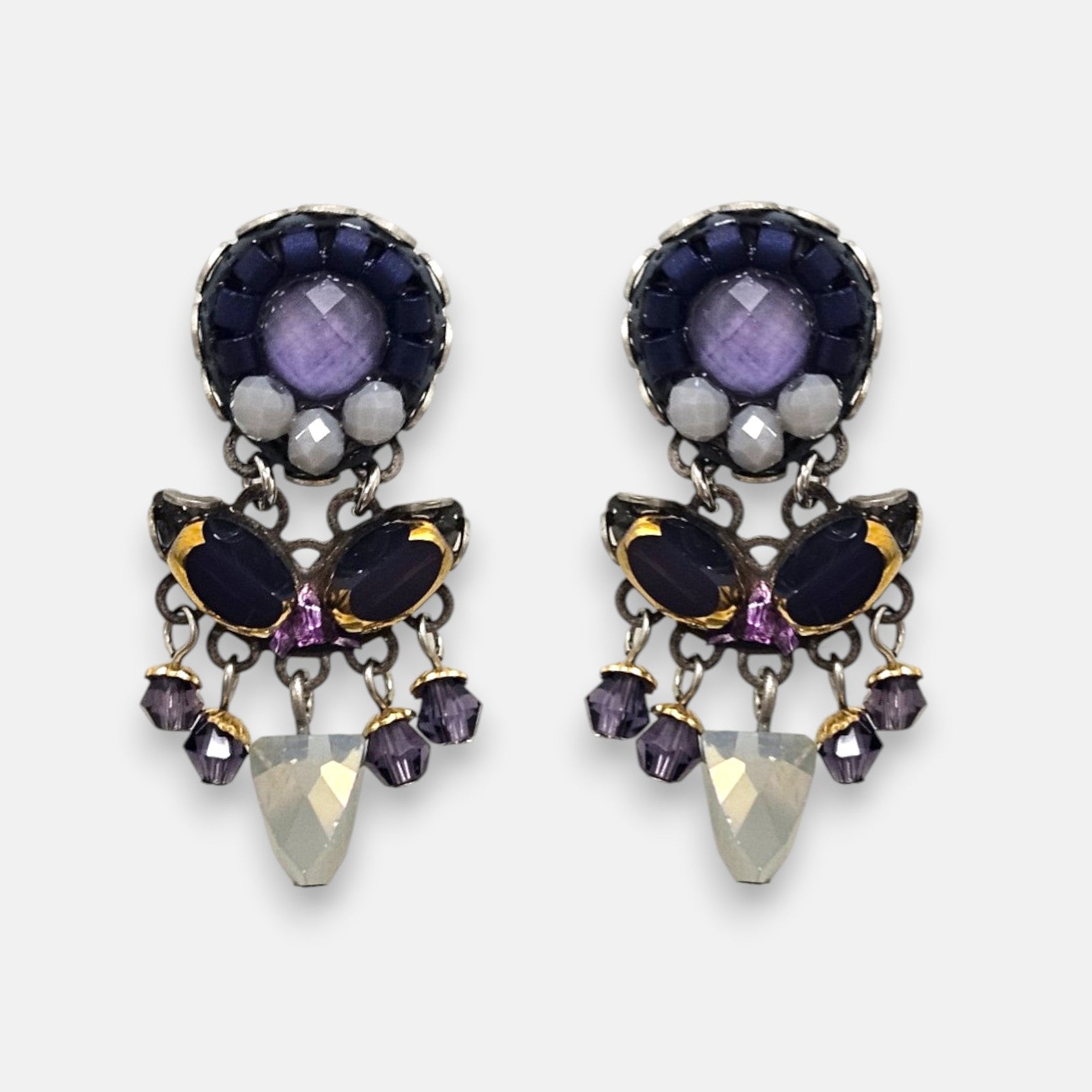 par de pendientes expuesto de frente sobre un fondo neutro, detallando su diseño simétrico de botón con cristales morados, azules y su colgante central opalescente.