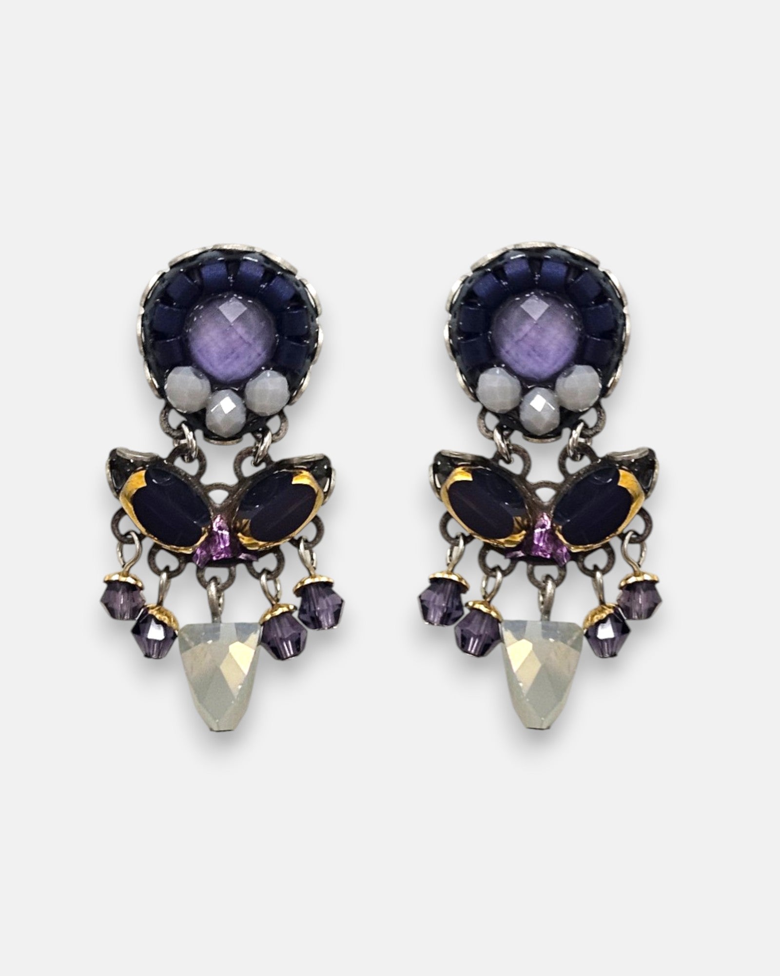 par de pendientes expuesto de frente sobre un fondo neutro, detallando su diseño simétrico de botón con cristales morados, azules y su colgante central opalescente.